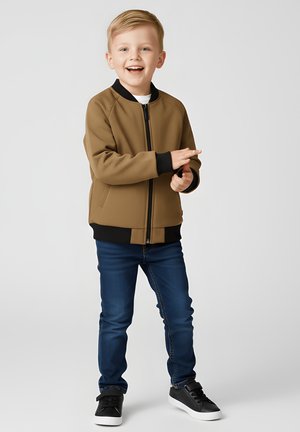 Brązowa kurtka bomberka z czarnymi detalami, z zamkiem błyskawicznym, w połączeniu z niebieskimi dżinsami i czarnymi trampkami. Dziecko stoi z zabawnym wyrazem twarzy.