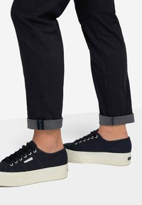 Sneakers in tela navy scuro con suola piatta bianca, punta rotonda, lacci navy e dettaglio in occhioletto argento. Indossati con pantaloni neri, risvoltati in grigio.
