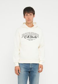 Cremefarvet hættetrøje med snore og en kengurulomme. Har et sort tekstlogo på forsiden. Parret med lyseblå jeans.