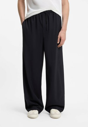 Osoba ubrana w luźne, szerokie czarne spodnie z elastycznym pasem i białe trampki, stojąca na jednolitym białym tle.