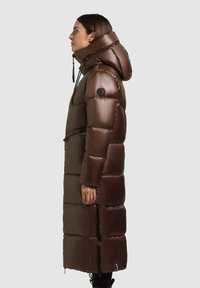 Barna párnázott hosszú kabát magas gallérral és kapucnival. Fényes felülettel, cipzárakkal és logóval ellátott patch-el a ujján.