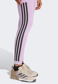 Leggings violets clairs avec trois bandes verticales noires le long de la jambe extérieure ; associés à des chaussures de sport beige avec des accents en maille et en matière solide.