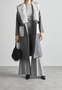 Woman wearing long gray coat with white fur trim and pockets, gray wide-leg pants, black pointed shoes, holding black furry handbag.