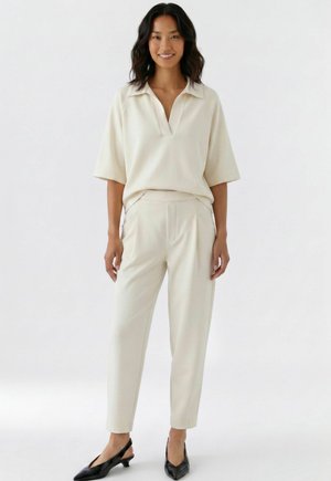 Femme souriante, portant un chemisier beige clair à manches courtes et un pantalon assorti avec des chaussures noires à talons bas et pointues, debout devant un fond blanc.