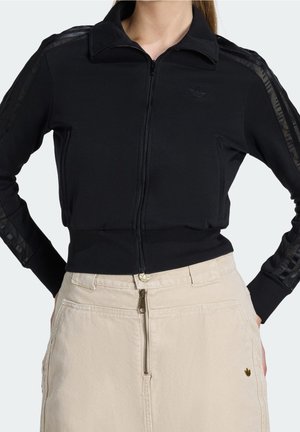 Femme portant une veste de survêtement noire zippée avec détails en mesh sur les manches et jupe beige avec fermeture éclair devant et emblème doré, les mains dans les poches.