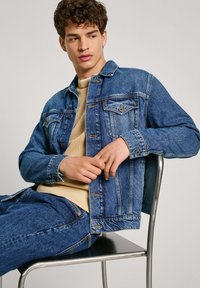 Chaqueta de mezclilla en azul intermedio con cuello clásico, cierres de botón y bolsillos en el pecho. Llevada sobre un suéter de textura beige claro.