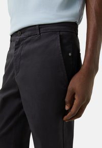 Dunkelblaue Hose aus glattem Stoff, mit seitlichen Taschen, einem Knopfverschluss und einem dezenten Markenakzent am Bund.