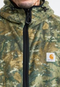 Jachetă din fleece cu un model de camuflaj, având nuanțe de verde și maro, fermoar frontal și o patch emblematică pe piept cu un design rotund portocaliu.