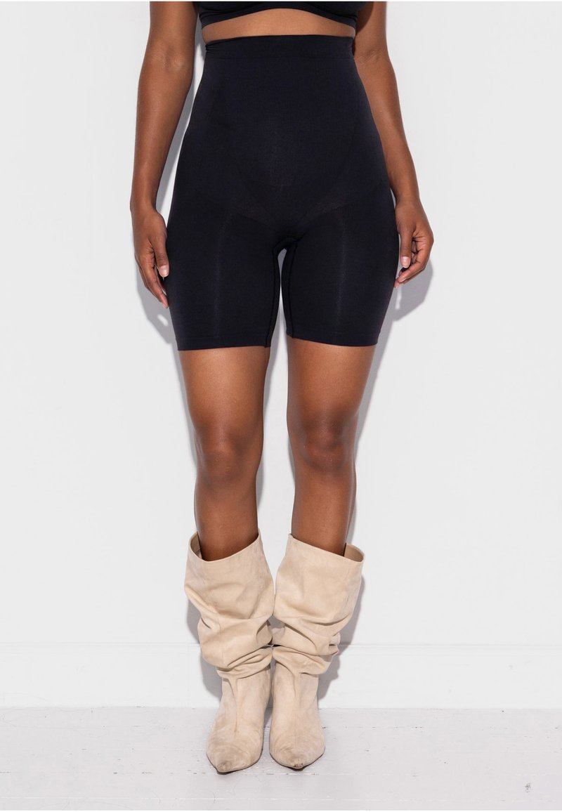 Nő, aki derékig érő fekete biciklis rövidnadrágot és laza, bézs színű térdig érő csizmát visel, egy fehér falnak támaszkodva áll.