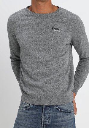 Homme portant un pull gris à col rond avec un petit logo noir sur la poitrine et un jean bleu, mains dans les poches, sur un fond uni.
