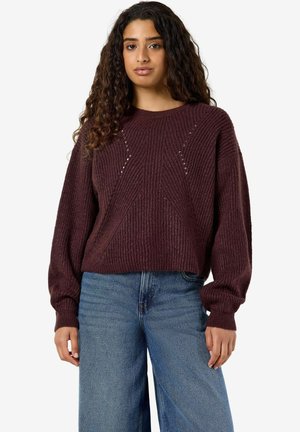Ung kvinde med langt krøllet hår iført en mørkelilla striktrøje og lyseblå jeans, stående mod en ensfarvet hvid baggrund.