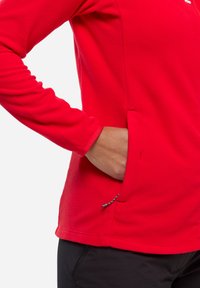 Haut long à manches rouges texturé avec une fermeture éclair et une poche latérale, présentant un détail avec cordon de serrage à l'ourlet. Le tissu semble léger et respirant.