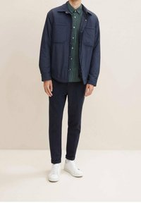 Giacca leggera navy sopra una camicia a scacchi verde, abbinata a pantaloni scuri e sneaker bianche. La giacca presenta due tasche sul petto.