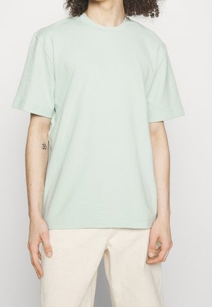 Persoon met krullend haar die een lichtgroen T-shirt met korte mouwen en lichtbeige broek draagt, en een kleine tatoeage op de linker onderarm toont.