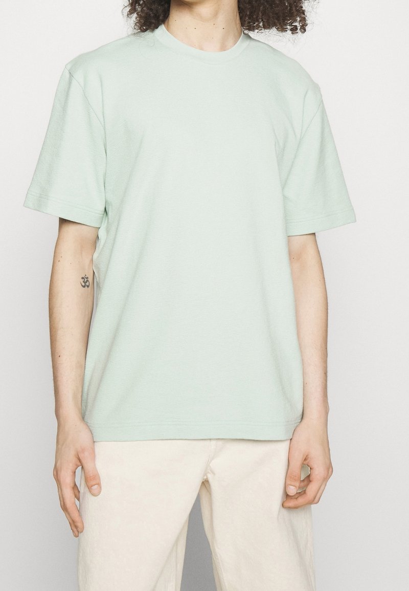 Personne aux cheveux bouclés portant un t-shirt à manches courtes vert clair et un pantalon beige clair, montrant un petit tatouage sur l'avant-bras gauche.