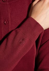 Maglione a maglia bordeaux con chiusura a bottoni, polsini a costine e un piccolo design ricamato sulla manica. Texture morbida e design aderente.