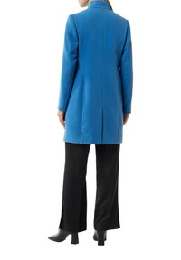 Blauer Wollmantel mit skrupulöser Silhouette, hohem Kragen und Rückenbelüftung. Kombiniert mit schwarzen weit geschnittenen Hosen und eleganten Knöchelstiefeln.