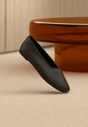 Fekete texturált, könnyen felvehető lapos cipő lebeg egy bézs szőnyeg felett, közel egy kerek, borostyánszínű asztallábhoz.