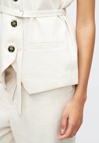 Gilet beige ajusté avec une texture en chevrons, doté de boutons noirs, d'une poche sur le côté et d'une ceinture à nouer pour l'ajustement.