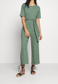 Groene, geribbelde jumpsuit met korte mouwen, voorzien van een geknoopte tailleband. Zwarte sandalen met bandjes maken de look compleet.