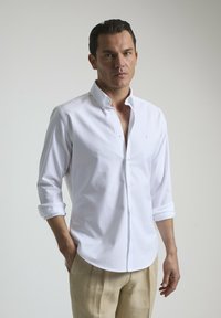 Camisa blanca de botones confeccionada en algodón, con cuello, mangas largas arremangadas hasta los codos y un sutil logo en el pecho.
