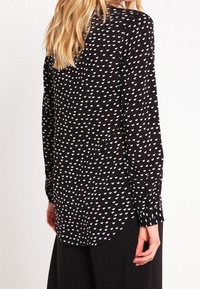 Svart blus med vitt hjärt-mönster, långa ärmar, rundad nederkant och knäppta manschetter. Slät tyg med lätt textur.