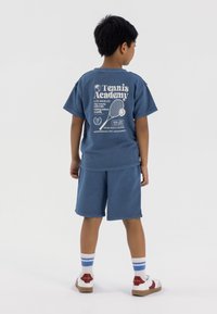 T-shirt di cotone blu con stampa grafica bianca e pantaloni corti coordinati. L'outfit presenta una vestibilità rilassata e maniche corte, abbinato a calzini a strisce e sneakers.