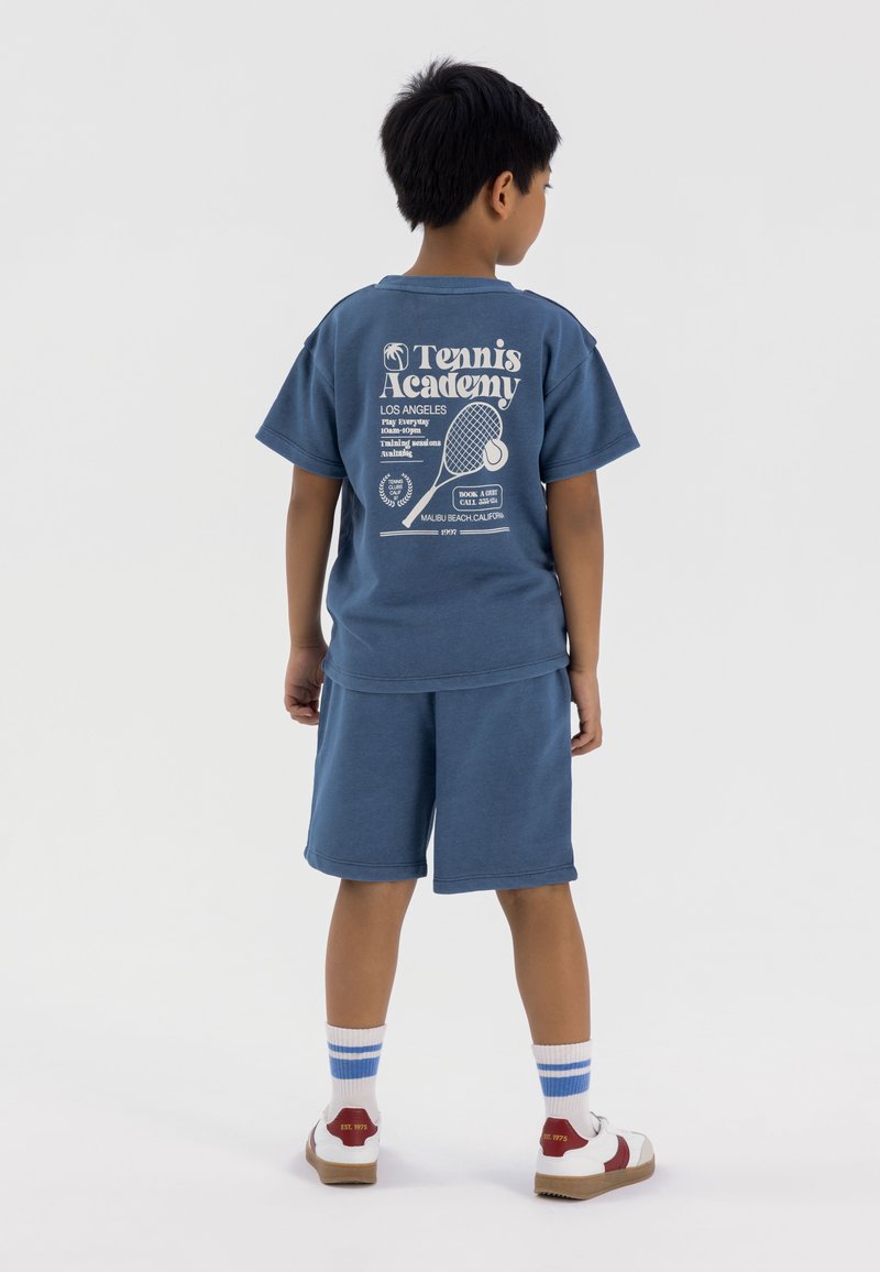 T-shirt di cotone blu con stampa grafica bianca e pantaloni corti coordinati. L'outfit presenta una vestibilità rilassata e maniche corte, abbinato a calzini a strisce e sneakers.