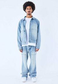 Giacca di jeans azzurro chiaro con dettagli a motivo diamante, abbinata a jeans larghi coordinati. T-shirt bianca sotto e sneakers bianche.