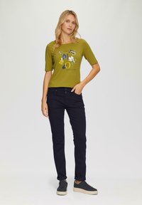 Camiseta de manga corta en verde oliva con estampado gráfico, combinada con pantalones ajustados en azul marino oscuro y zapatillas grises con suela texturizada.
