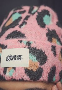 Rozā adīta cepure ar leoparda rakstiem melnā, tirkīza un oranžā krāsā. Ar baltu etiķeti, uz kuras ir "eleanor bowmer" logo.