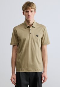 Polo tričko s krátkym rukávom z olivovo zeleného bavlneného materiálu, s klasickým golierom, zapínaním na tri gombíky a nášivkou s logom na ľavej strane hrudníka.