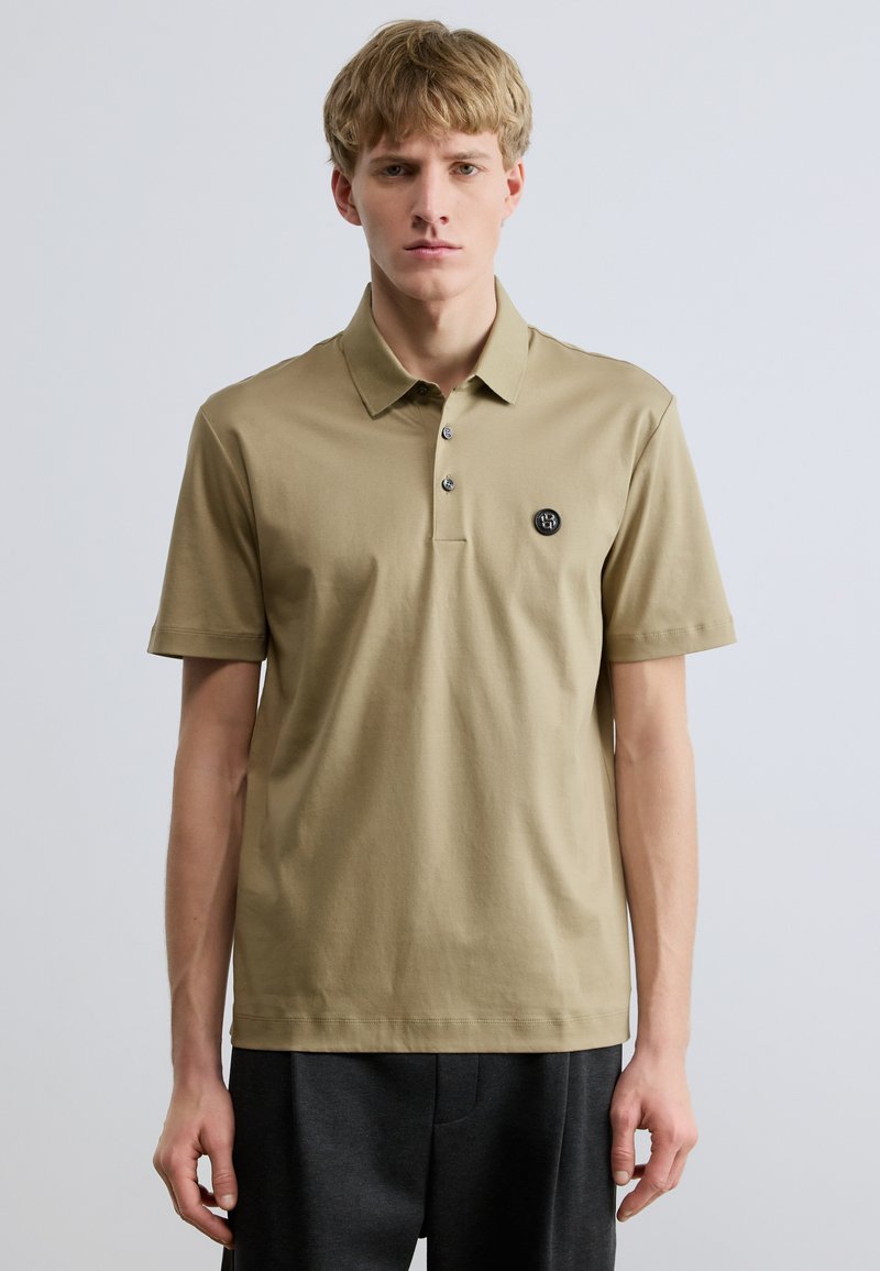 Polo tričko s krátkym rukávom z olivovo zeleného bavlneného materiálu, s klasickým golierom, zapínaním na tri gombíky a nášivkou s logom na ľavej strane hrudníka.