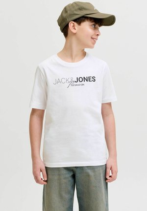 Jongen draagt een wit Jack & Jones Premium T-shirt, olijfgroene pet en een blauwe jeans, kijkt opzij en lacht tegen een effen achtergrond.