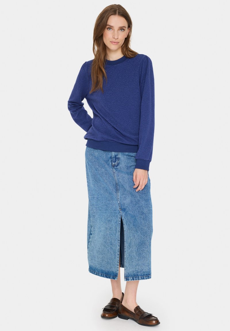 Marineblaue Sweatshirt mit Struktur und langen Ärmeln, kombiniert mit einem hochgeschnittenen Jeansrock mit Schlitz vorne und braunen Slippern.