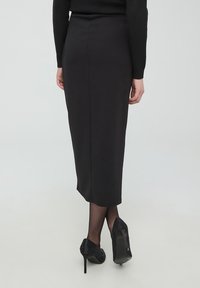 Gonna midi nera, aderente, con una texture liscia e uno spacco posteriore. Abbinata a scarpe col tacco nere con design borchiato.