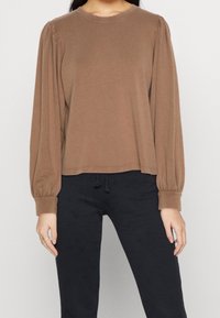Maglietta marrone a maniche lunghe con colletto rotondo, presenta maniche arricciate, una texture morbida e una vestibilità rilassata, abbinata a pantaloni neri con cordoncino.