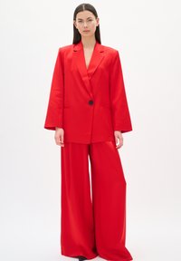 Roter Blazer mit einem einzelnen schwarzen Knopf, weiten Ärmeln und passenden weiten Hosen. Glattes Material mit einer taillierten Passform, das ein stimmiges Outfit kreiert.
