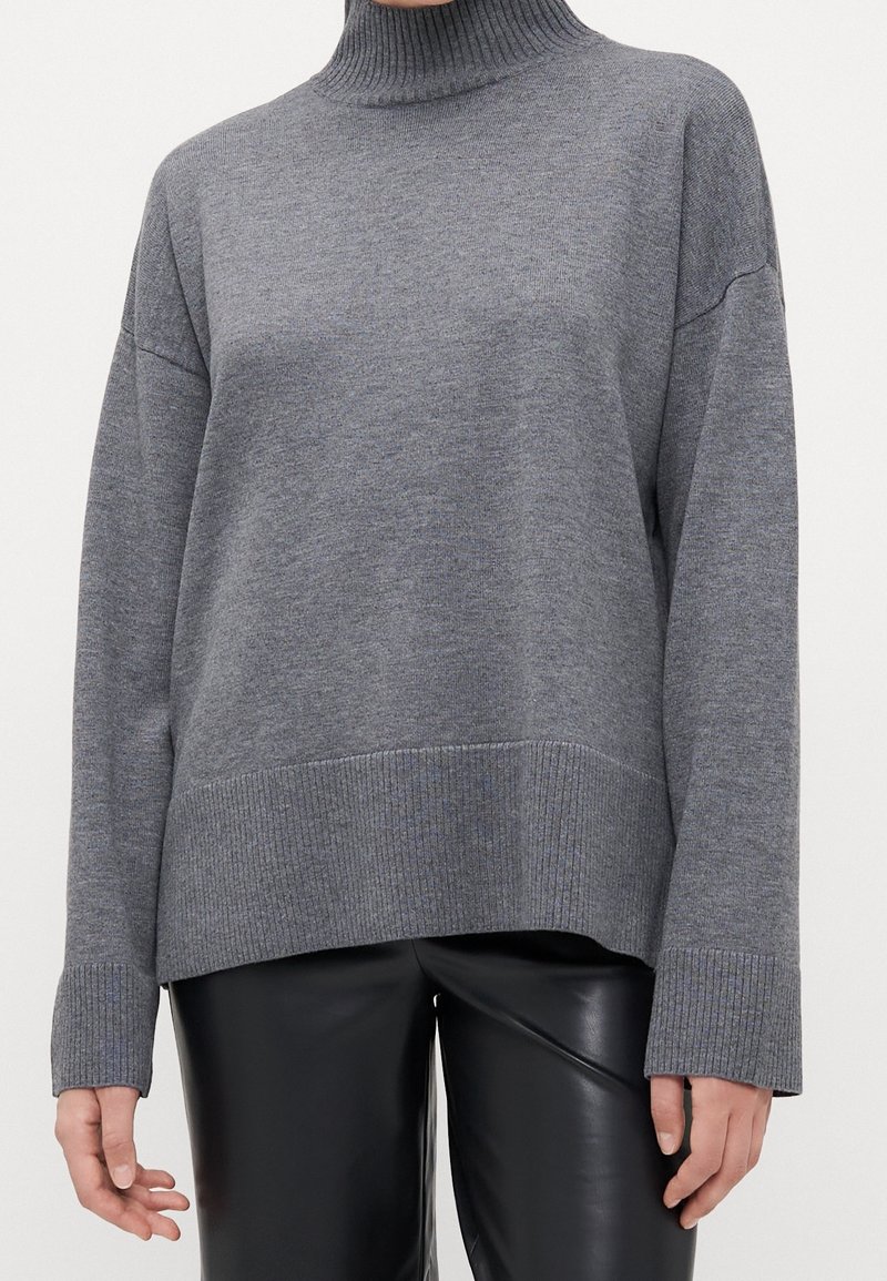 Personne portant un pull col roulé gris ample associé à un pantalon en cuir noir sur un fond uni.