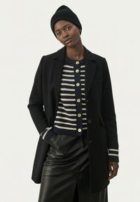 Svart kappa med slag, över en marinblå och vit randig cardigan med guldknappar. Svart läderskjorta och en ribbad svart mössa.