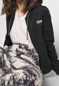 Felpa nera con zip, logo dorato e lacci con motivo. Indossata sopra una maglietta chiara, abbinata a pantaloni bianchi e neri con motivo.