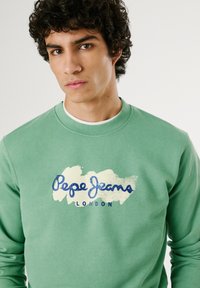 Grüner Baumwollpullover mit Rundhalsausschnitt, mit dem Aufdruck "Pepe Jeans London" in Blau und Creme auf der Vorderseite. Strukturierter Stoff.