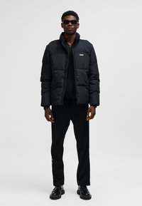 Sort dunjakke med lynlåslukning, der har en høj krave og logo-patch. Modellen bærer sorte bukser og sko, suppleret med solbriller.