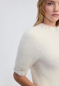 Witte, pluizige trui met korte mouwen en een hoge halslijn, versierd met decoratieve parels en zilveren accenten aan de zoom. Zachte textuur is duidelijk zichtbaar.