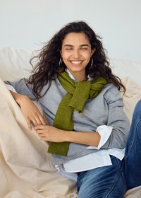 Jeune femme aux longs cheveux bouclés, portant un pull gris et une écharpe verte, assise confortablement sur un canapé beige, souriant les yeux fermés.