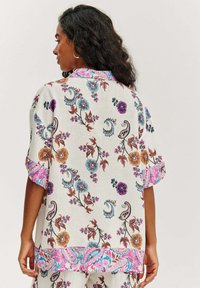 Bílá květinová košile s vícebarevnými vzory v modré, fialové a oranžové barvě, s růžovým akcentem na rukávech a spodním lemu.
