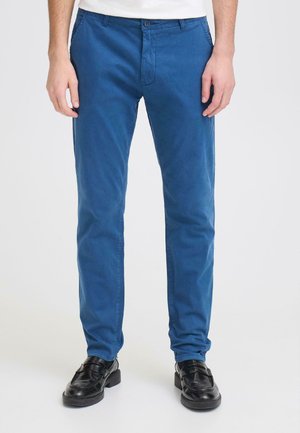 Man draagt slim fit blauwe broek en zwarte leren loafers, staand tegen een effen lichte achtergrond.