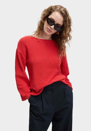 Rode gebreide trui met een brede halslijn en lange mouwen, gecombineerd met donkere geplooide broeken. Model draagt zwarte zonnebril en creolen.