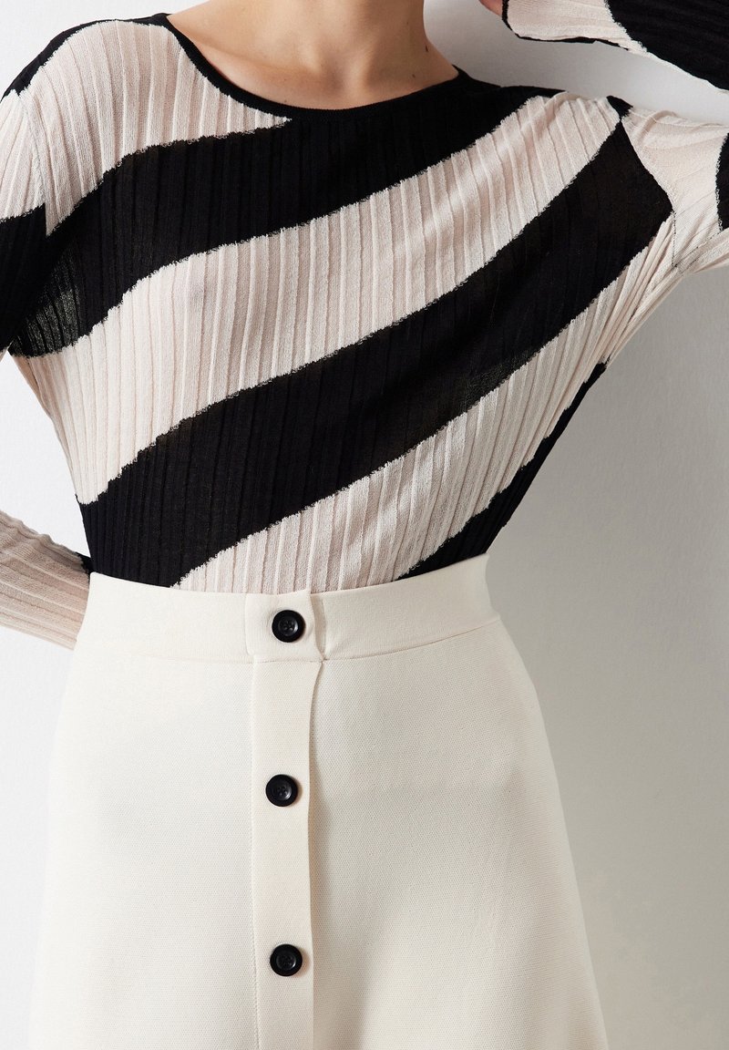 Femme portant un haut côtelé à manches longues rayé en diagonale noir et blanc, rentré dans une jupe crème taille haute avec des boutons noirs.