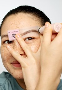 Øyenbrynsmal fra Revolution holdt mot et ansikt. Har klart plastikk med rosa etikett og applikatorrør i myk rosa, som fremhever forming av øyenbryn.